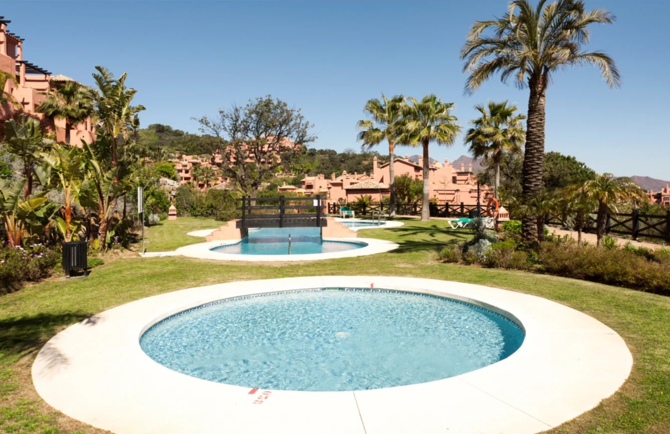 Zona de ocio ajardinada de una urbanización con césped, flores, palmeras y varias piscinas redondas