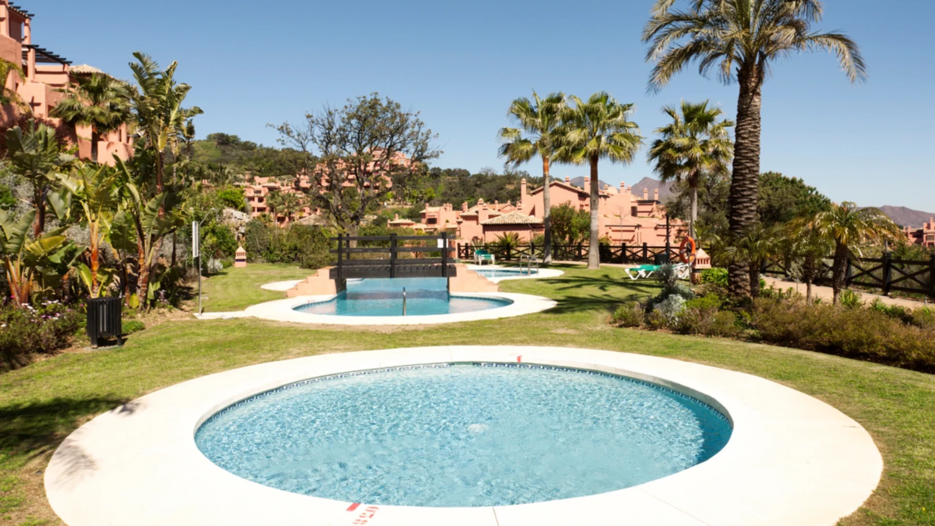 Zona de ocio ajardinada de una urbanización con césped, flores, palmeras y varias piscinas redondas